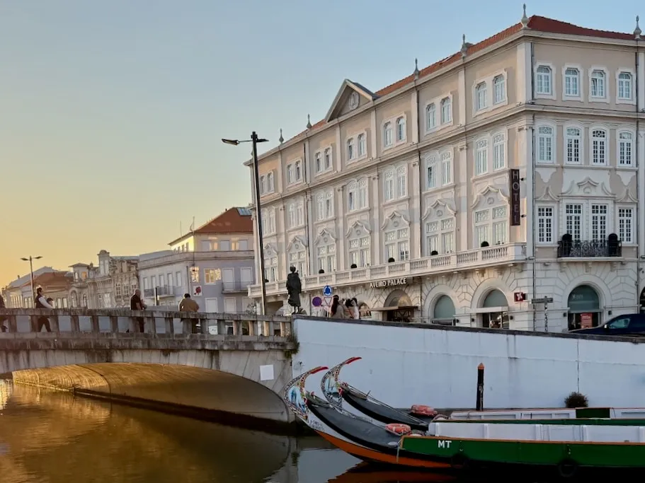Traditional moliceiro boats moored on a canal in central Aveiro at sunset, beside a stone bridge and an ornate pale building.